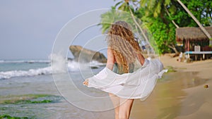 Curly-haired woman in green swimsuit, white sarong, strolls along sandy shore, invites viewers to tropical beach getaway