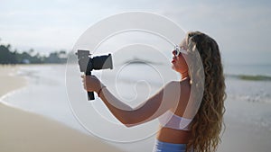 Curly-haired woman records with DSLR on sunny beach, waves crashing. Content creator captures coastal scenery, uses