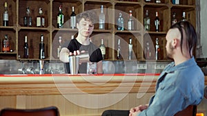 Curly-haired young Caucasian man pouring tasty alcohol drink from shaker in glass with ice for client. Barkeeper serving