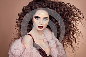 Curly hairstyle. Fashionable elegant woman with makeup and blowing healthy long hair posing in pink fur coat isolated on beige ba