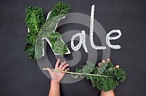 Curly-leaf kale cabbage inside letter K shaped plate, chalk inscription KALE on blackboard, child`s hands holding fresh greens.