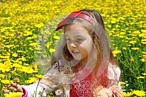 Curly little girl with a red bow in her hair. Girl with a dol on a green meadow among yellow flowers