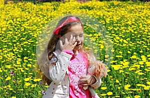 Curly little girl with a red bow in her hair. Girl with a dol on a green meadow among yellow flowers