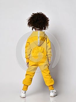 Curly mulatto dark-skinned kid girl in warm blue sports jumpsuit with hood and sneakers stands back to camera