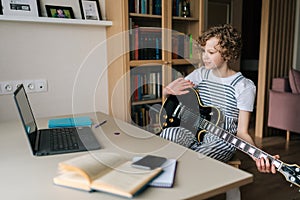 Curly preteen girl learning to play the guitar in virtual meeting together with friend or teacher in video conference.