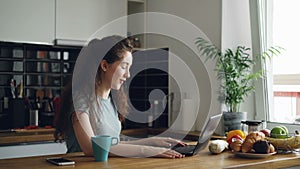 Curly pretty positive caucasian woman sitting at table skyping on laptop, she is talking sitting in kitchen near window