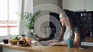 Curly pretty positive caucasian woman sitting at table skyping on laptop, she is talking sitting in kitchen near window