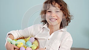 Curly red-haired child in a white shirt with a figure of an Easter bunny and a set of Easter eggs in a basket. A boy