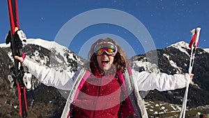 Curly skier rejoices in the snow against the backdrop of mountains. Woman in mirror glasses, ski suit and skis in hand