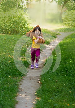 Curly smiling girl in a yellow T-shirt holds a large brown teddy bear on her neck and runs along the path