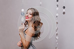 Curly stylish lady in shining dress coquettishly looks into camera on white background with silver glitter.