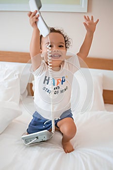 Curly toddler boy playing with a wired telephone on the bed