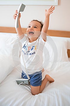 Curly toddler boy playing with a wired telephone on the bed