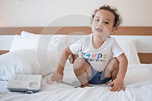 Curly toddler boy playing with a wired telephone on the bed