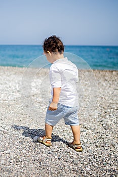 Curly toddler boy plays on the pebble beach looking to blue sea