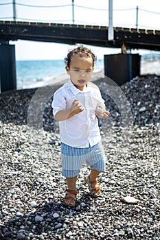 Curly toddler boy plays on the pebble beach near blue sea