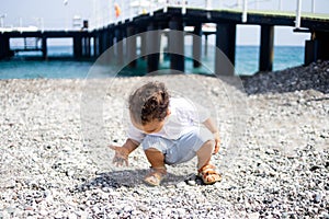 Curly toddler boy plays on the pebble beach near blue sea
