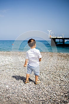 Curly toddler boy plays on the pebble beach near blue sea