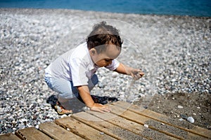 Curly toddler boy plays on the pebble beach near blue sea