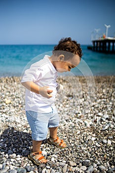 Curly toddler boy plays on the pebble beach near blue sea