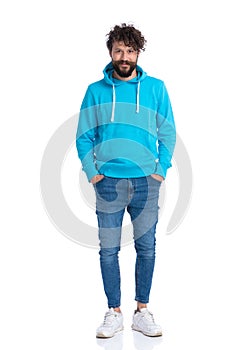 Curly unshaved man with blue sweatshirt holding hands in pockets