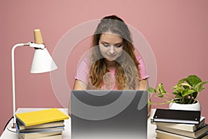 Curly woman in a pink t-shirt using laptop computer while sitting at a desktop in a pink wall studio.