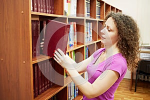 Curly woman takes large book from bookshelf in