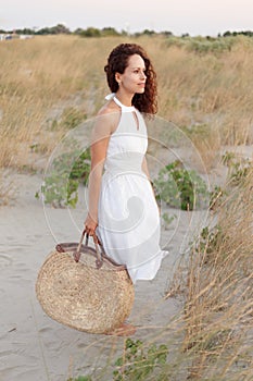 Curly woman in white dress with bag of straw going to the beach at the sunset