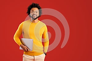 Curly young African-American male entrepreneur stands in studio isolated on red and carrying trendy laptop. Cheerful