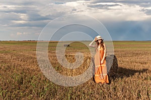 Curly young rural teenager girl stands near a bundle of hay in a sundress and hat on a harvested wheat field with a backdrop of a
