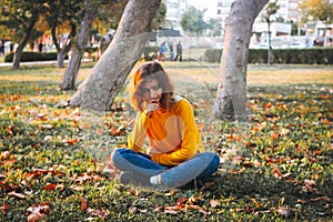 Curly young girl in yellow sweater and jeans sitting on fall grass with dry leaves. Autumn mood