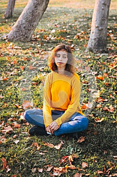 Curly young girl in yellow sweater and jeans sitting on fall grass with dry leaves. Autumn mood