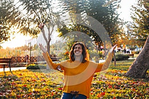 Curly young girl in yellow sweater and jeans sitting on fall grass and throws up dry leaves