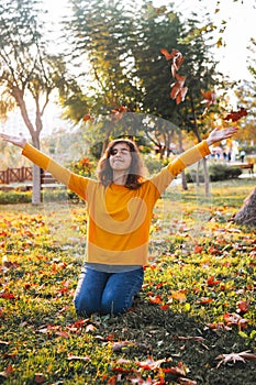 Curly young girl in yellow sweater and jeans sitting on fall grass and throws up dry leaves