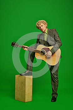 Curly young man in a suit playing guitar against green studio background. Poster for live music event
