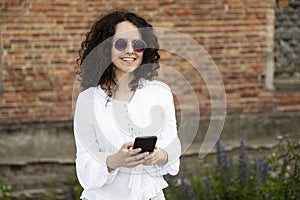 Curly young woman, looking at the camera, holding a cell phone, against a building with red bricks background.