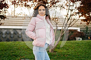 Curly young woman in pink jacket walks in the park