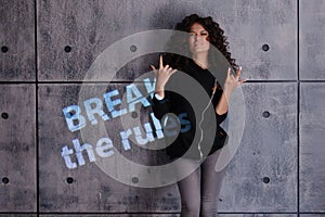 Curly young woman in a strong, boxing pose against a concrete wall background. Colored light sign Break the rules.