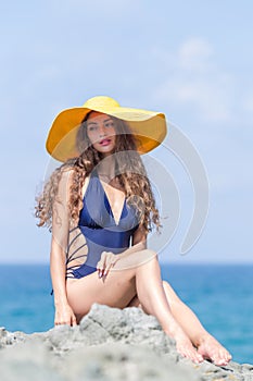 Curly young woman in yellow brim board hat sits on rock against sea