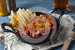 Currywurst with french fries in black pan on craft paper served with beer on wooden table.