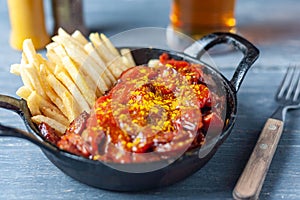 Currywurst with french fries in black pan served with beer on wooden table.