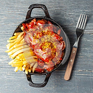 Currywurst with french fries in black pan on wooden table.