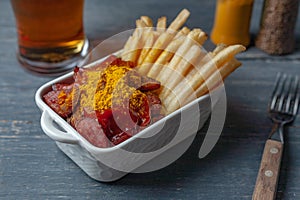 Currywurst with french fries in white bowl on wooden table served with beer.