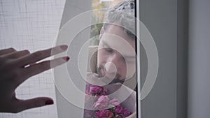 Curtain opening on the glass door and adult Caucasian gray-haired man standing behind it with a bouquet of violet roses