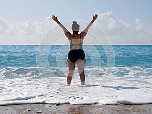 A curvaceous woman in a black bathing suit stands in the sea, raising her hands