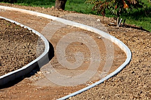 Curved concrete curb at local sidewalk construction site filled with gravel and small grain sand surrounded with grass and trees