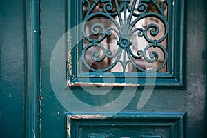 Curved ornate gratings of old wooden door painted in aquamarine green color. Blurry reflection in door window glass behind swirls