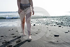 Curvy adolescent walking in the beach in the sunset in the wet black sand and waves on the sea. Caucasian woman in bikini at the