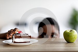 Curvy African American woman choosing between cake and apple, peeking at food from under table, free space