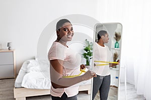 Curvy Afro woman measuring waist with tape near mirror, satisfied with weight loss result, achieving slimming success